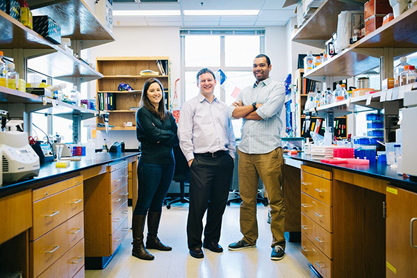 Two students smile while standing alongside their instructor in a lab.